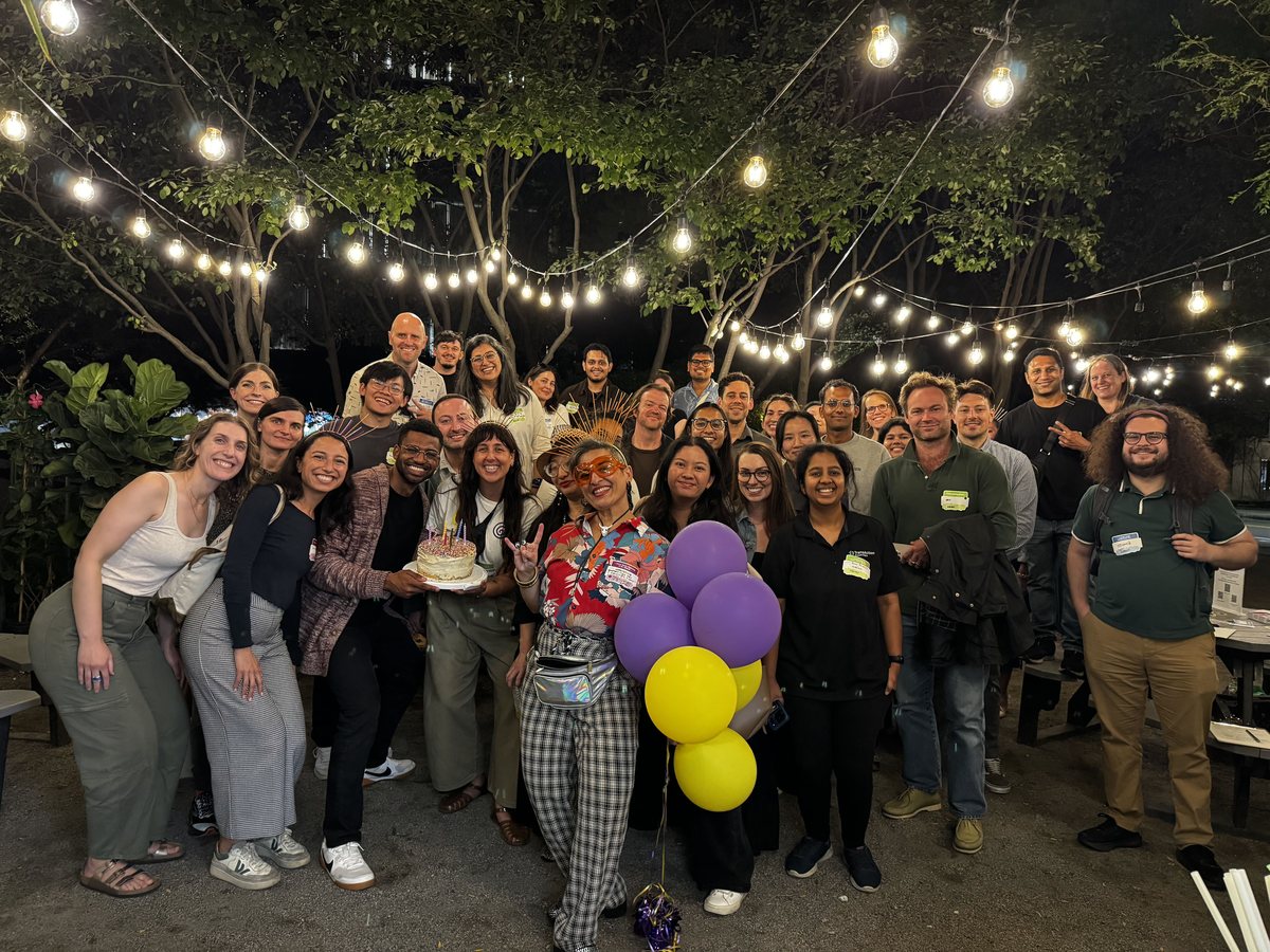 A large group of Generalist World members at an evening meetup under string lights, holding a cake and balloons