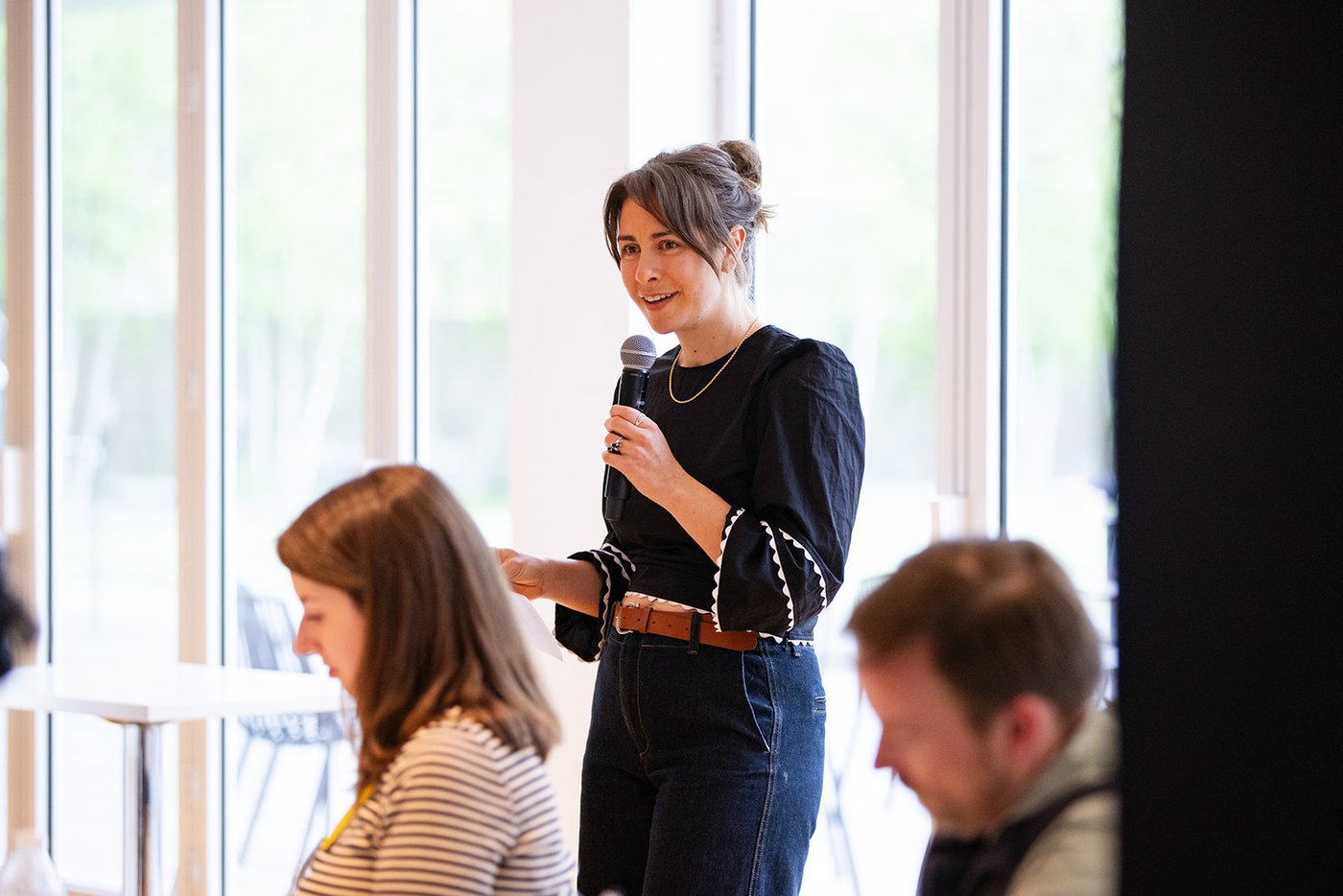 Milly speaking into a microphone in a bright room with floor-to-ceiling windows, audience in the foreground