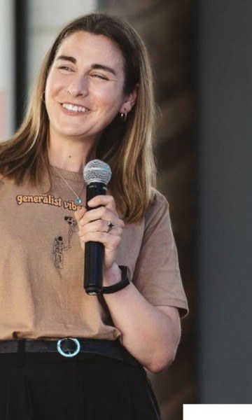 Milly on stage with a microphone, laughing, in a generalist vibes T-shirt