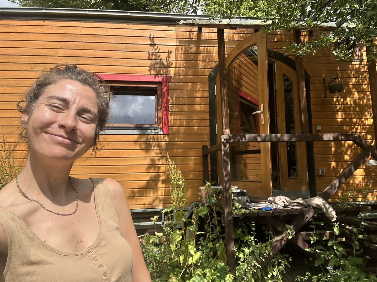 Milly smiling outside her wooden tiny home, with an arched door and red window frames